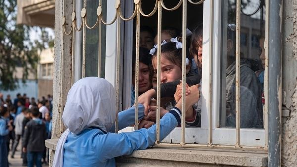 Okulun parmaklıklı penceresinin ardındaki çocuklara dışarıdan el uzatan, başörtülü, mavi üniformalı bir öğrenci